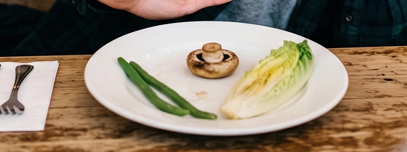A hand pushing away a plate with two string beans, one mushroom, one lettuce leaf